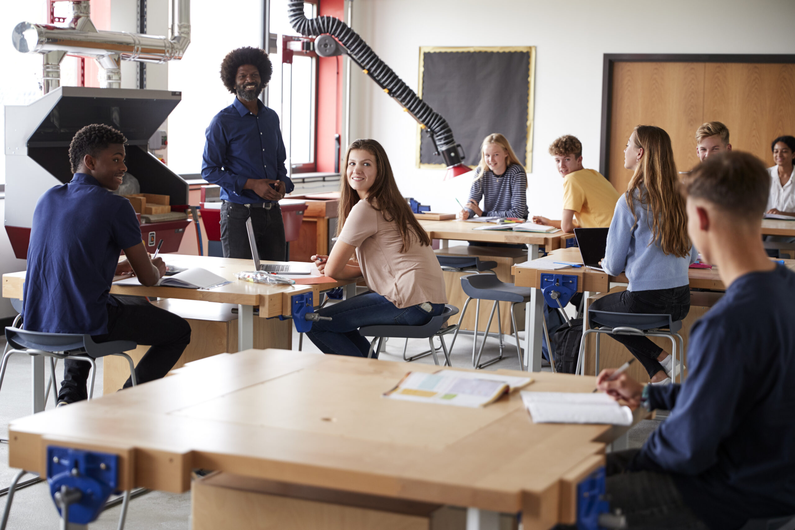 Class Of High School Students Sitting At Work Benches Listening To Teacher In Design And Technology Lesson